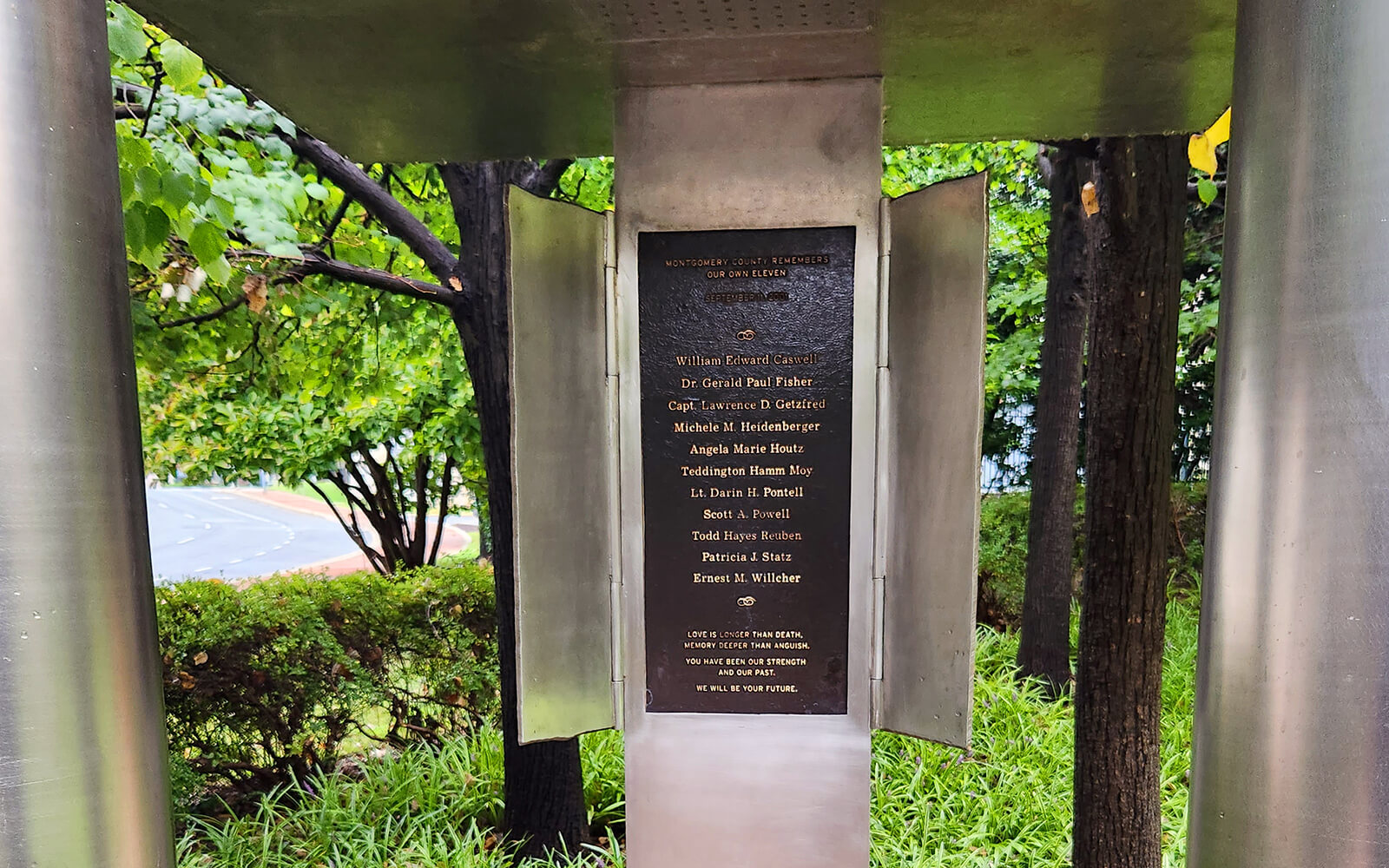 A metal memorial plaque with several engraved names is mounted outdoors among green trees and bushes.