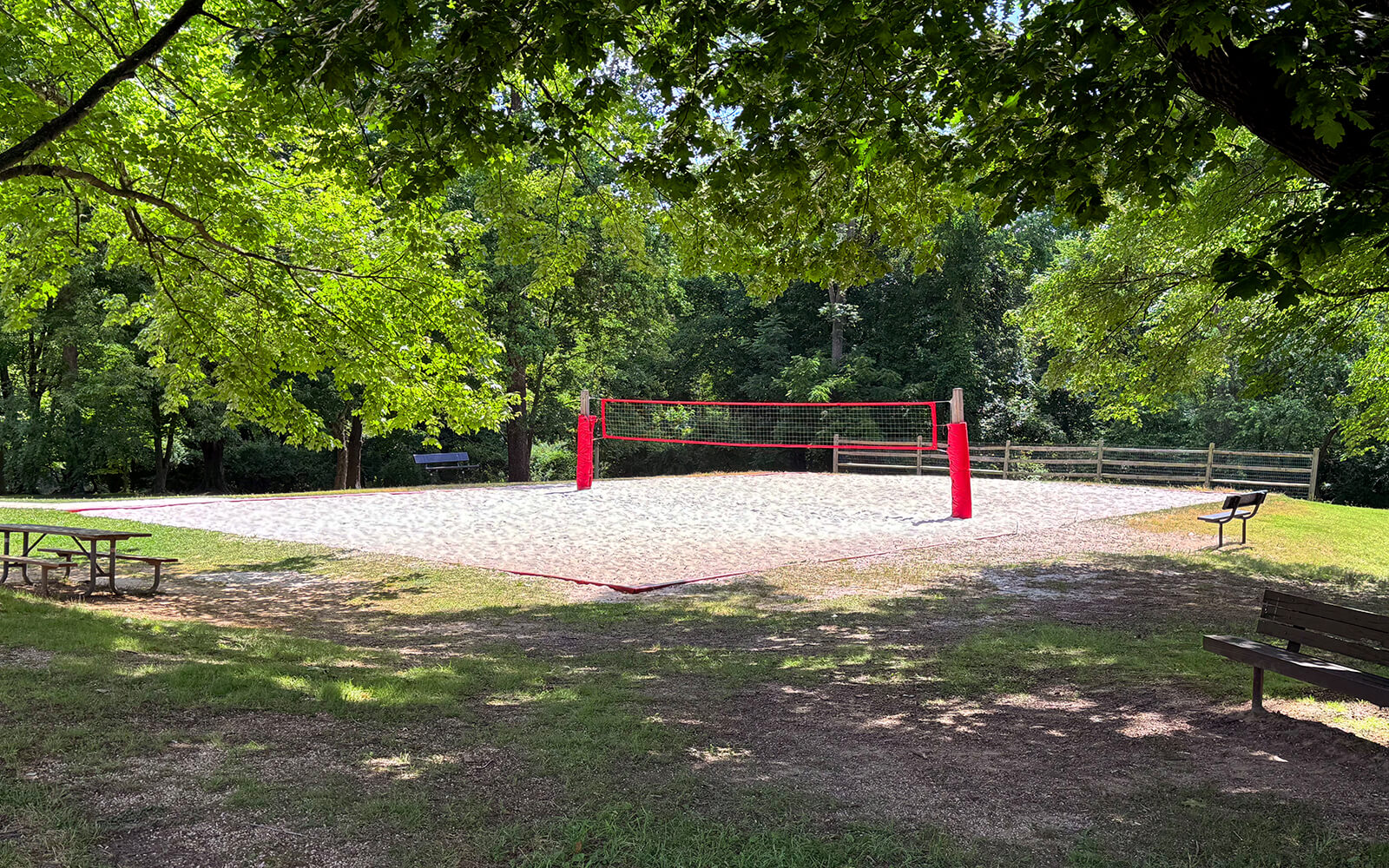A sand volleyball court with red poles is set in a park surrounded by trees, with picnic tables and benches nearby.