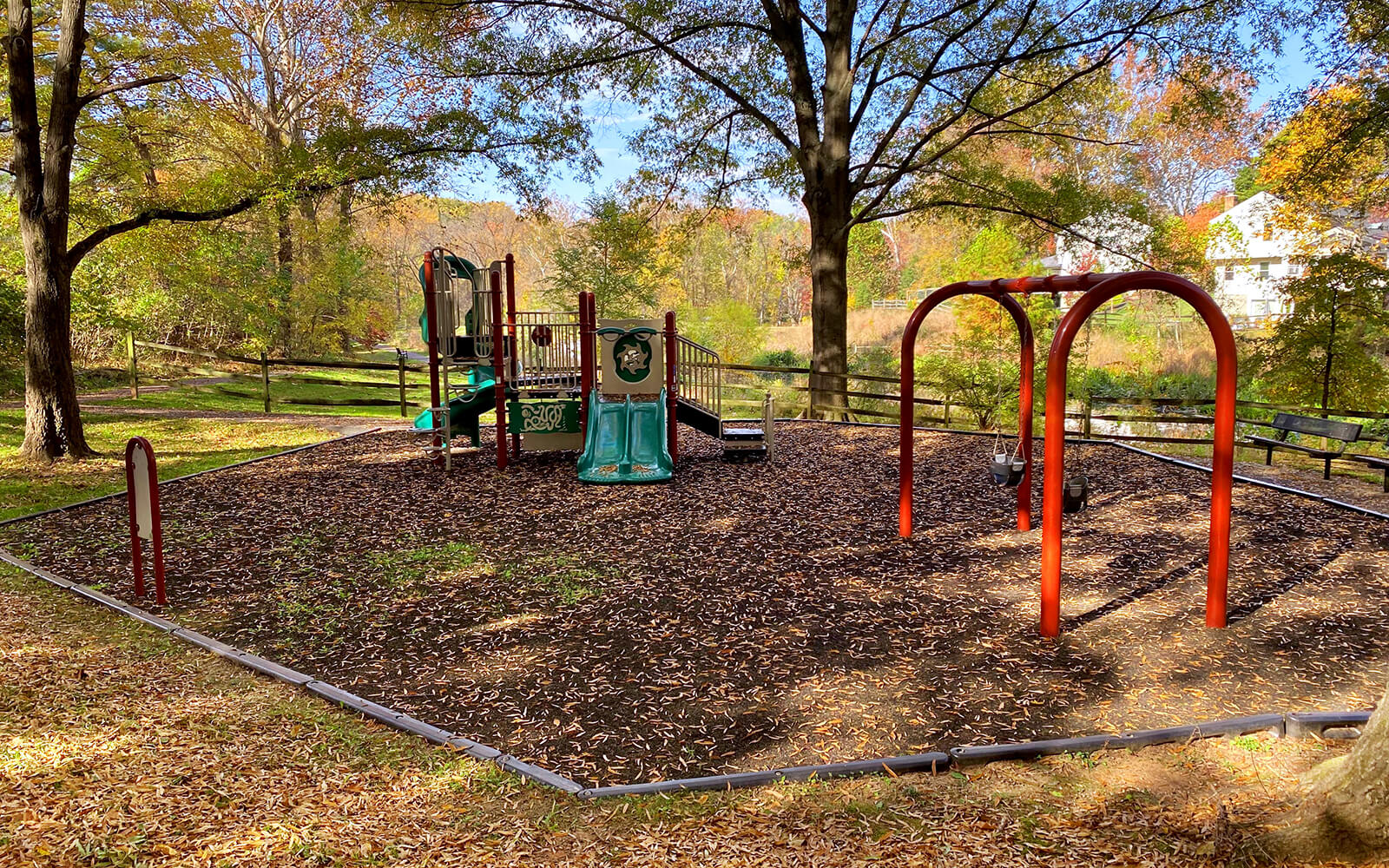 A small playground with slides, climbing equipment, and swings stands on a leaf-covered area surrounded by trees and a wooden fence.