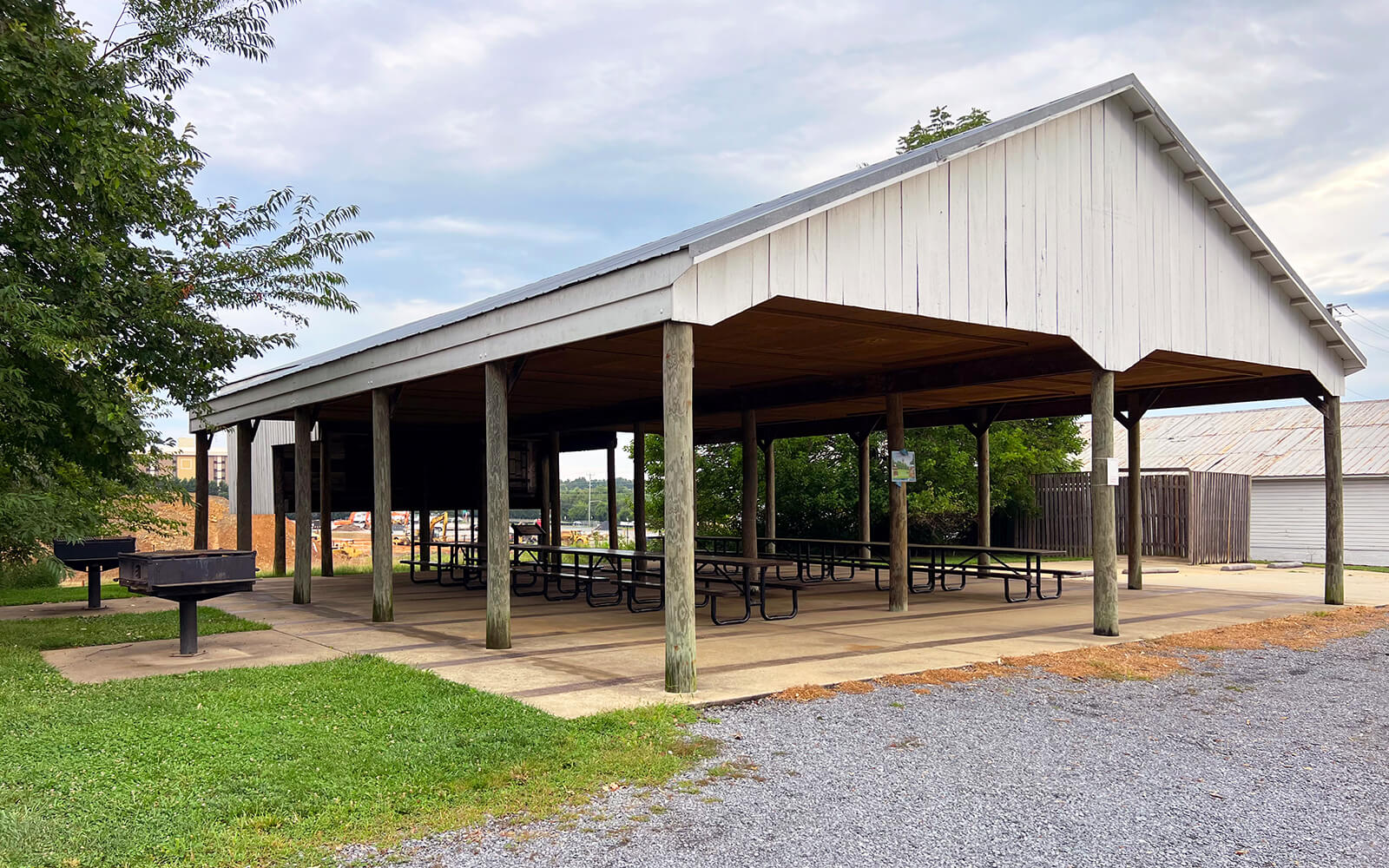 A large, open-sided picnic shelter with multiple tables underneath, situated on a concrete pad near a grassy area and gravel, with a charcoal grill nearby.