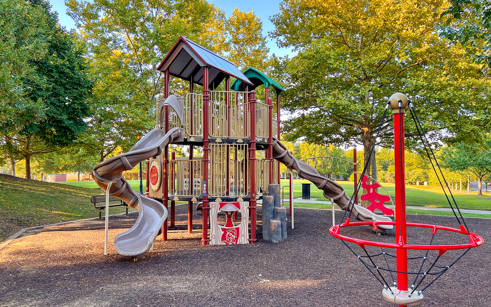 A modern playground with climbing structures, slides, and a rope spinner, surrounded by trees and green grass on a sunny day.