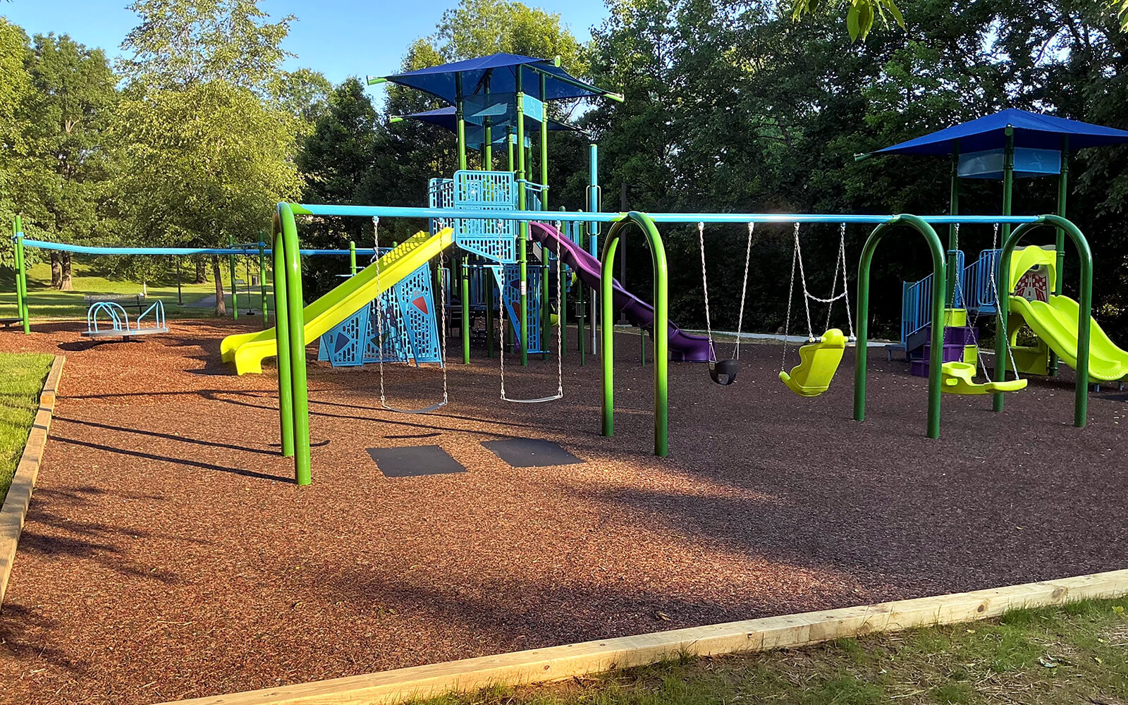 Outdoor playground with slides, swings, climbing structures, and protective ground cover, surrounded by trees and greenery.