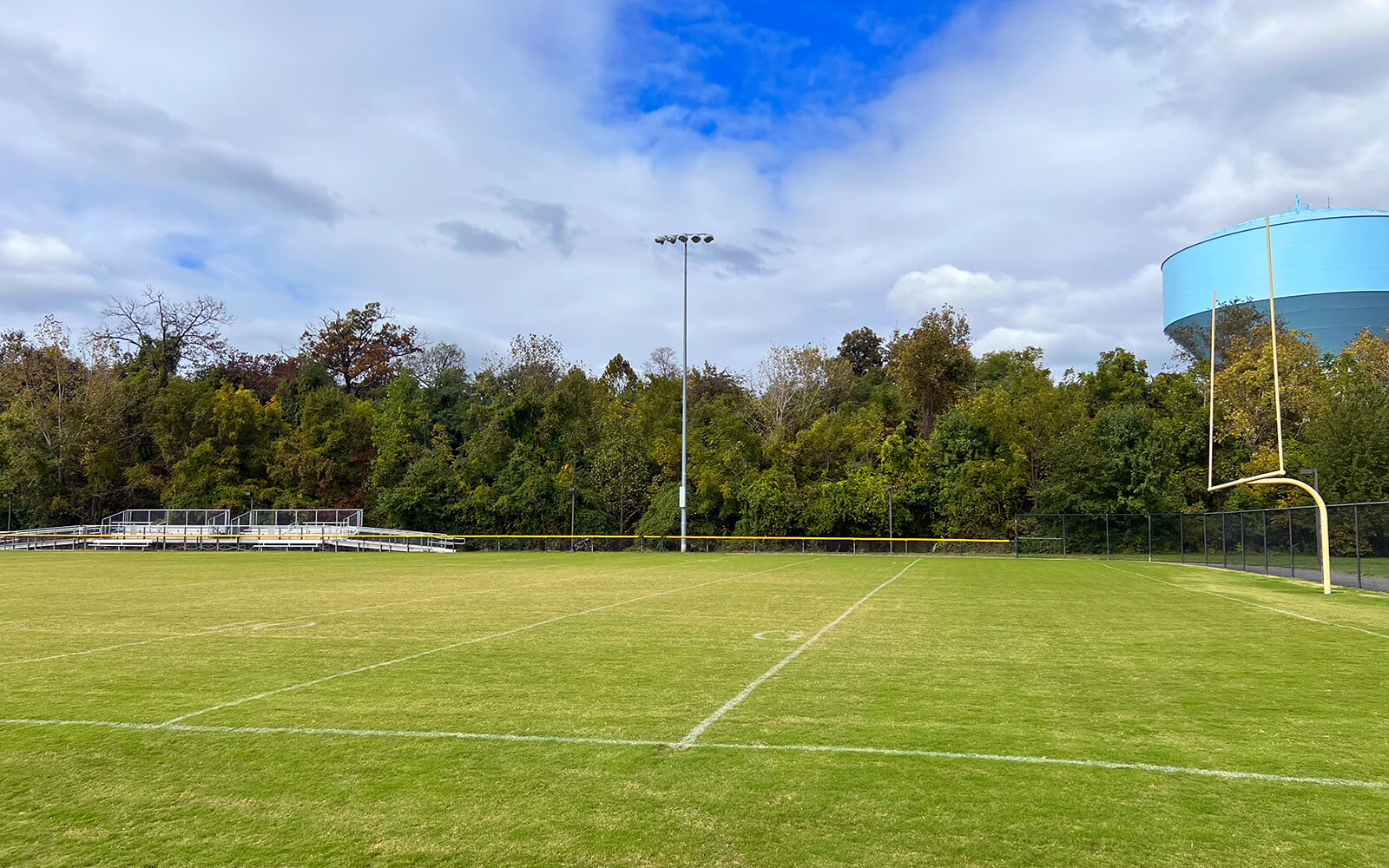 A grassy football field with white boundary lines, empty metal bleachers, a goalpost, and trees in the background under a partly cloudy sky.