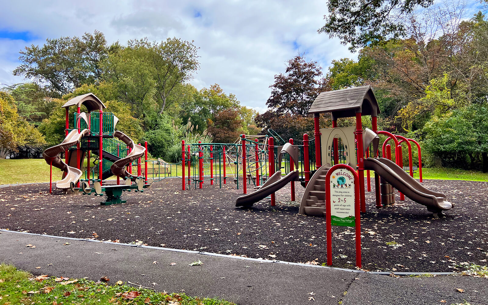 A playground with multiple slides, climbing structures, and monkey bars, set on a mulch surface, surrounded by trees and grass under a partly cloudy sky.