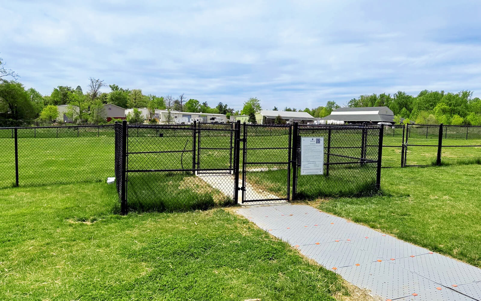 A fenced dog park entrance with a gate, sign, and paved walkway, surrounded by green grass and trees, with houses in the background under a partly cloudy sky.