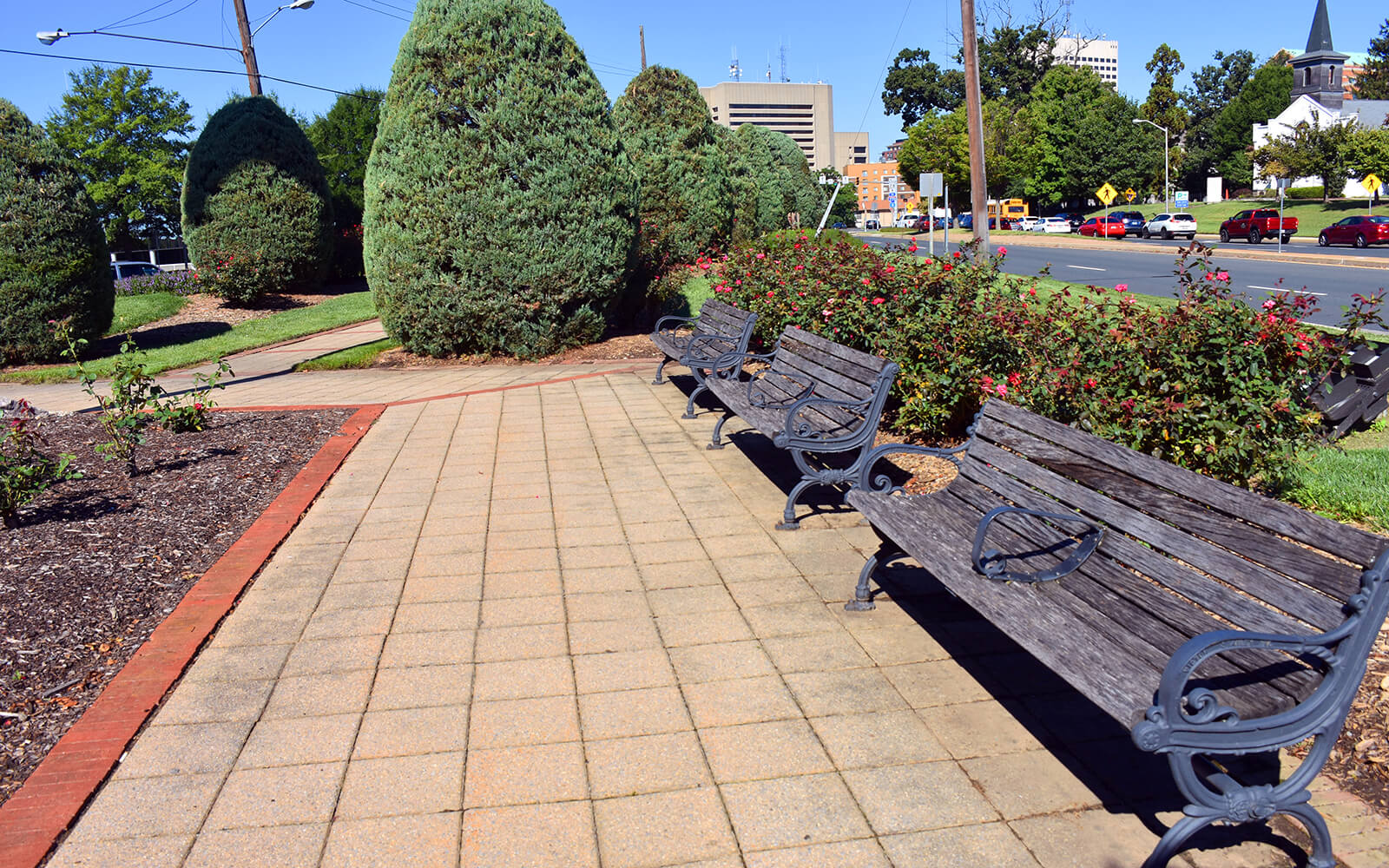 A paved walkway with empty wooden benches, bordered by bushes and trees, runs alongside a road with visible cars and buildings in the background.