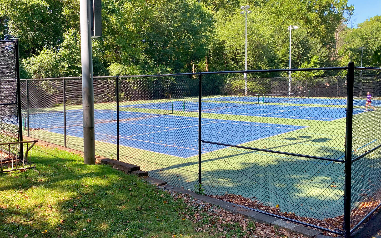 Two blue and green outdoor tennis courts are enclosed by a black chain-link fence with trees and greenery surrounding the area. A person stands on the far court.