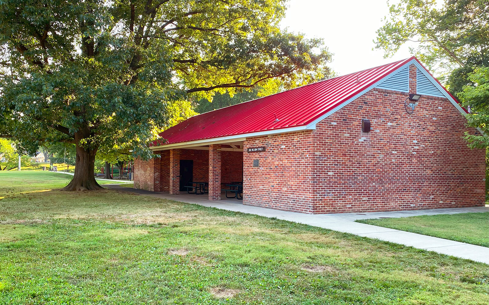 A brick pavilion with a red roof and covered picnic tables stands beside a large tree in a grassy park area.