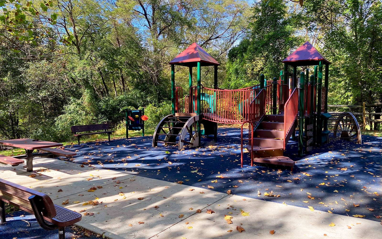 Outdoor playground with slides, climbing structures, benches, and picnic tables, surrounded by trees and scattered fallen leaves on a sunny day.