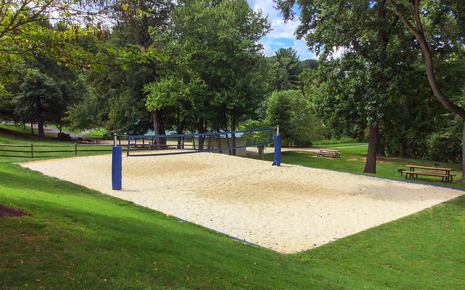 A sand volleyball court with two blue poles is surrounded by grass, picnic tables, and trees in a park setting.