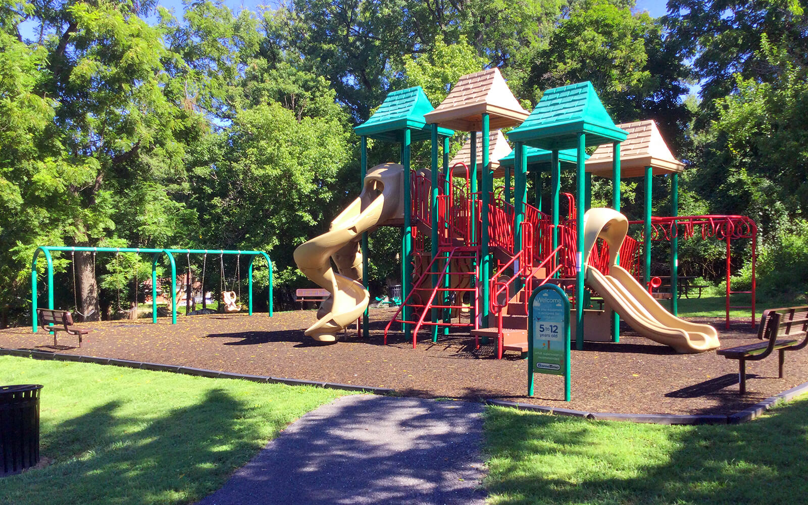 Outdoor playground with slides, swings, and climbing structures surrounded by trees and grass; benches and a trash can nearby under clear daylight.