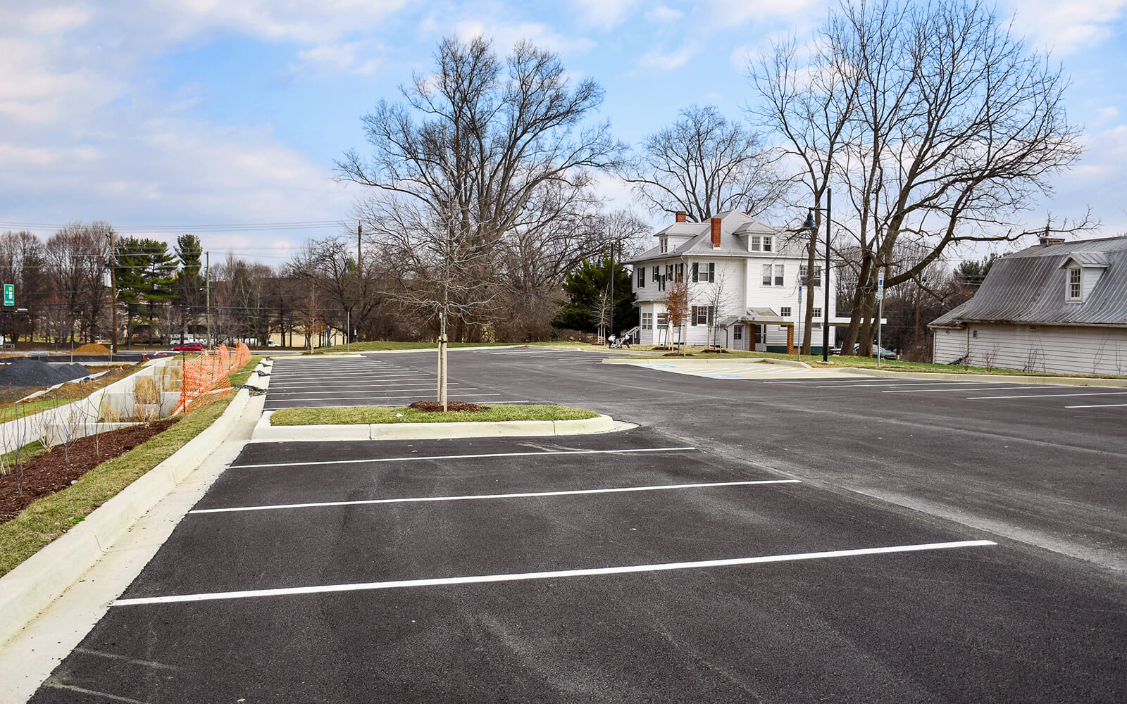 An empty paved parking lot with marked spaces is shown next to a large white house and bare trees under a partly cloudy sky.