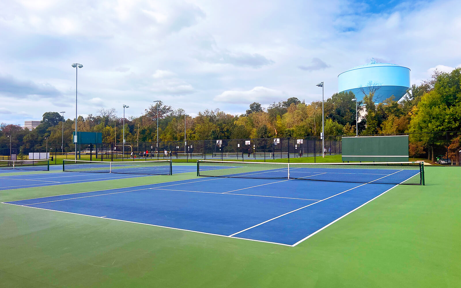 Outdoor tennis courts with blue and green surfaces, surrounded by fencing, trees, and a large blue water tower in the background under a partly cloudy sky.