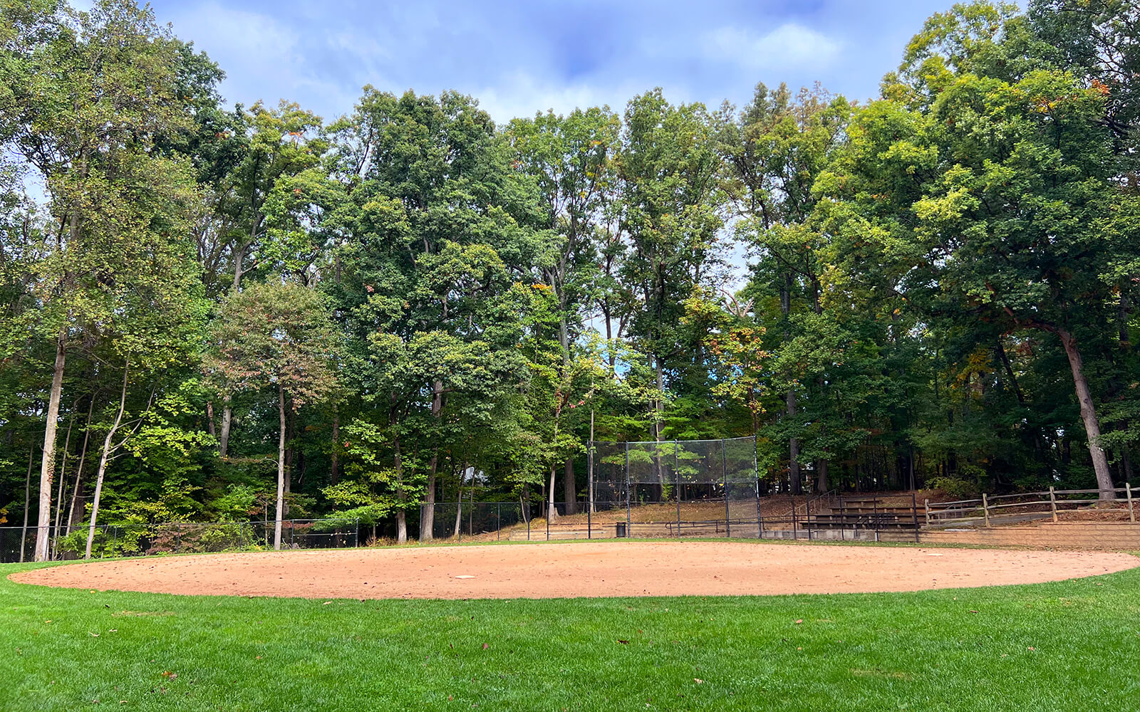 A baseball field with a dirt infield, surrounded by green grass, trees, and a wooden fence, under a partly cloudy sky.