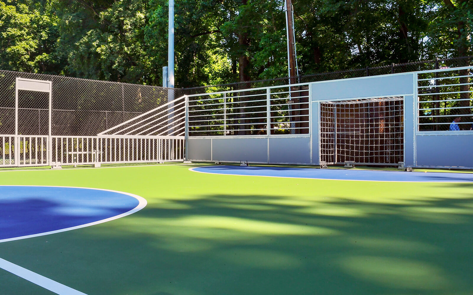 Outdoor futsal court with blue and green flooring, metal fencing, and two goals, surrounded by trees and sunlight creating shadows on the surface.