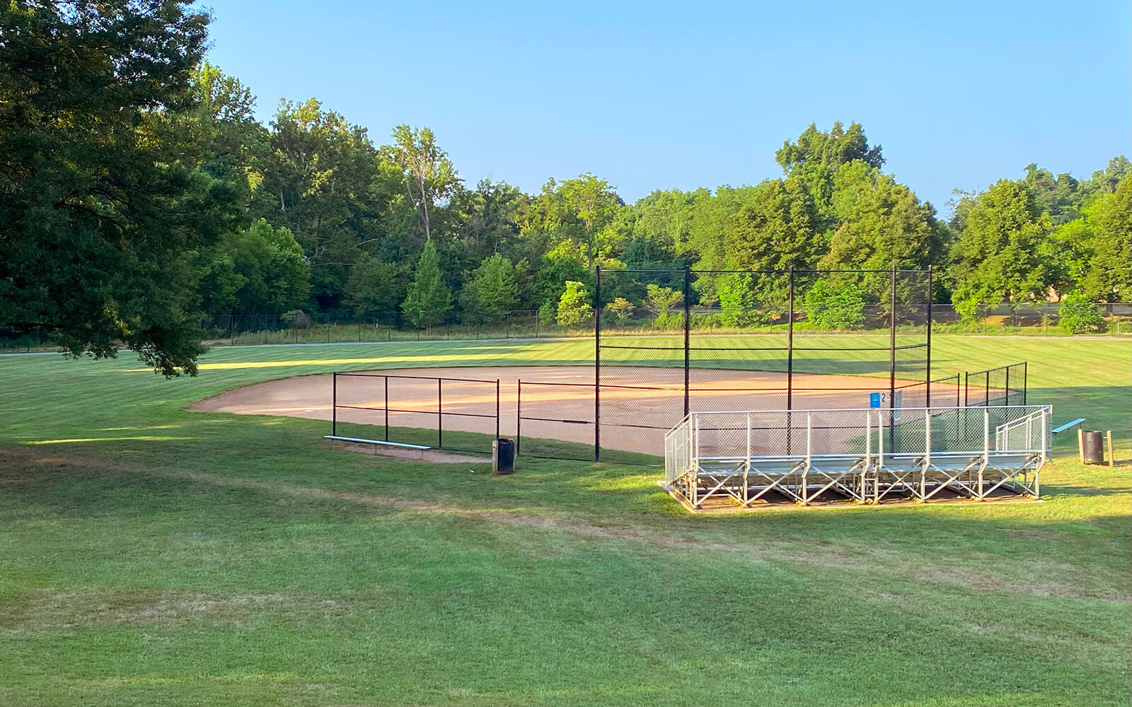 An empty baseball field with metal bleachers, surrounded by grass and trees under a clear blue sky.