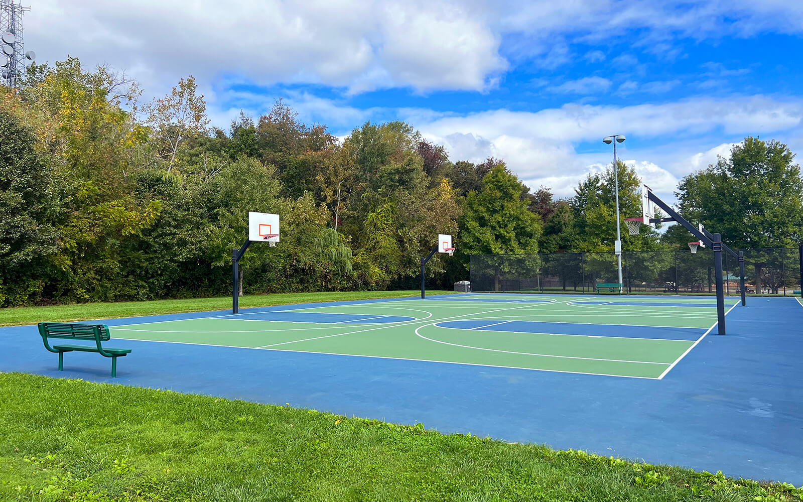 Outdoor basketball courts with blue and green surfaces, surrounded by trees and grass, under a partly cloudy sky; a green bench is in the foreground.