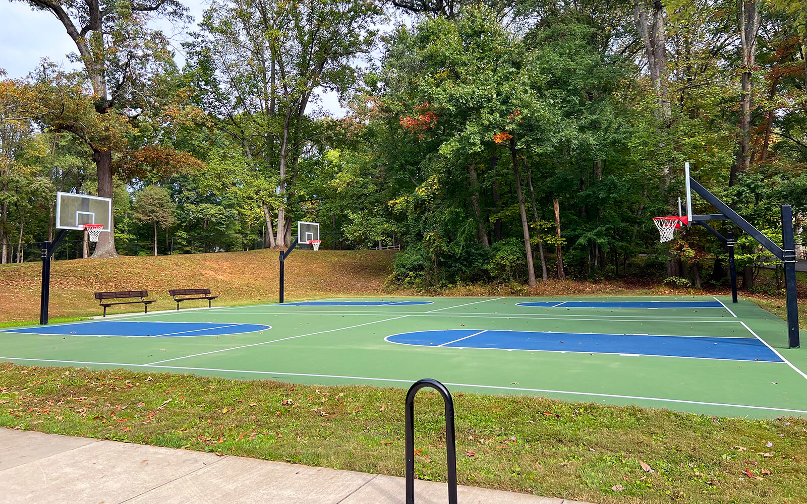 Outdoor basketball court with four hoops, surrounded by trees and benches on a clear day. The court has green and blue markings.
