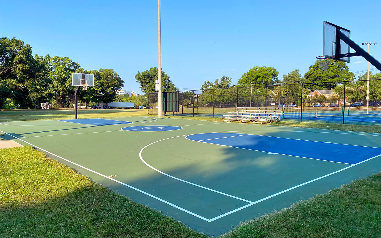 Outdoor basketball court with two hoops, surrounded by grass, trees, and a fence; metal bleachers and tennis courts are visible in the background under a clear blue sky.