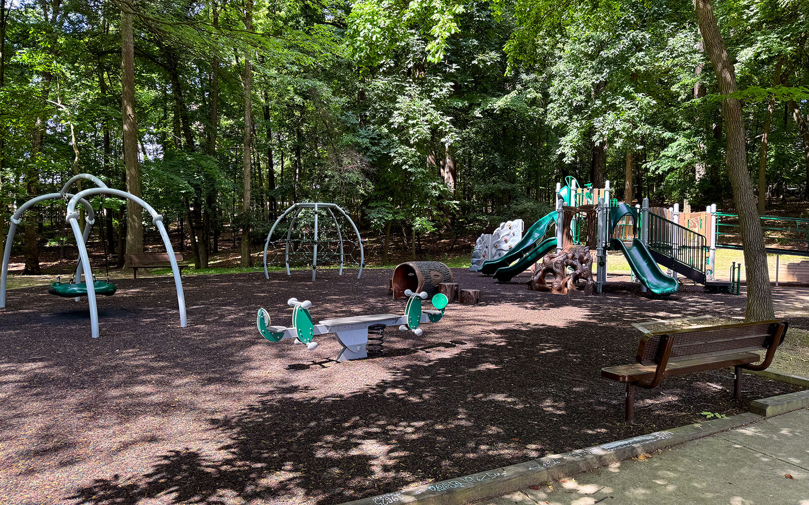 A playground with swings, climbing structures, slides, seesaws, benches, and trees providing shade in the background.