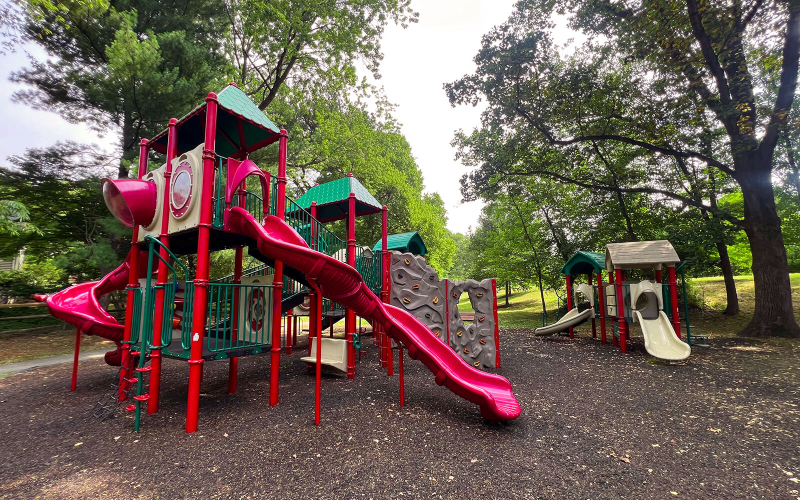 A playground with red and green play structures, including slides, climbing walls, and bridges, surrounded by trees and mulch ground covering.