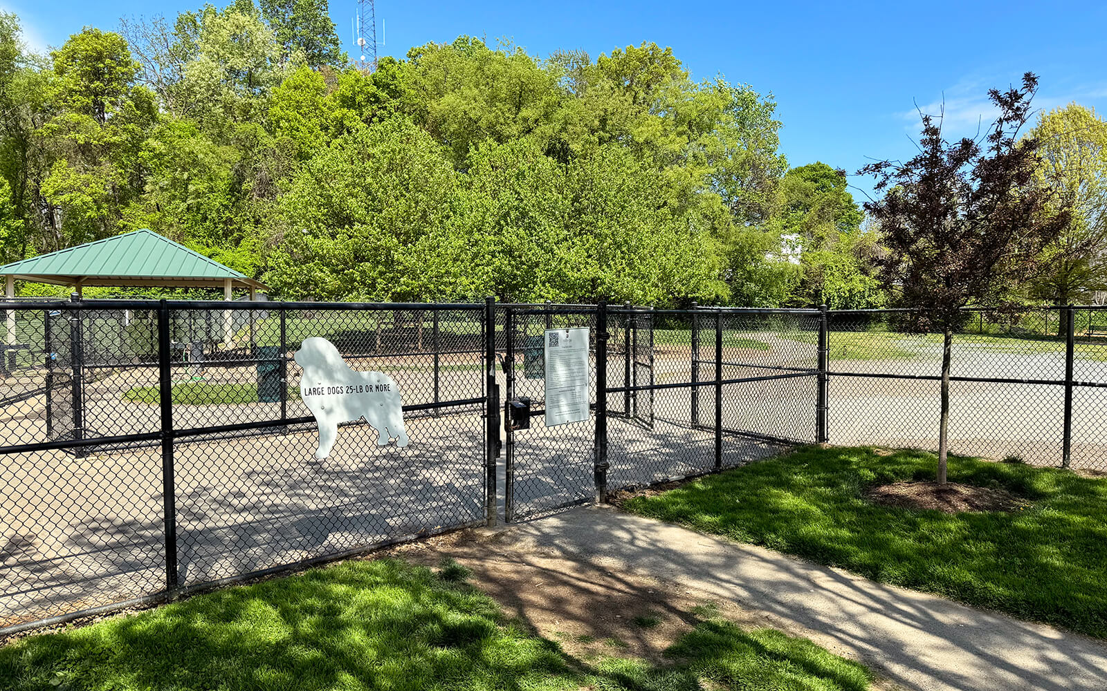 A fenced dog park with a white dog-shaped sign on the gate, surrounded by green trees and grass under a clear blue sky.