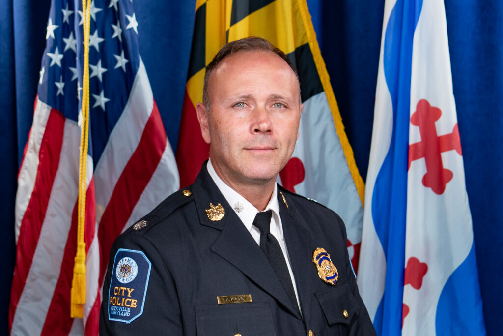 A police officer in dress uniform stands in front of the American flag, the Maryland state flag, and another blue and white flag.