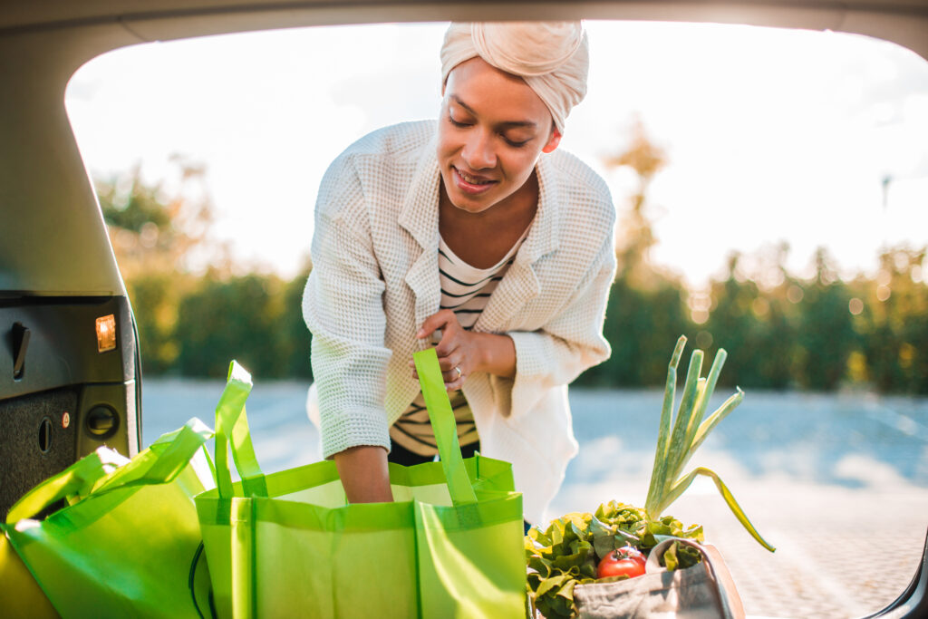 A person wearing a headwrap and striped shirt unloads green reusable shopping bags filled with vegetables from a car trunk.