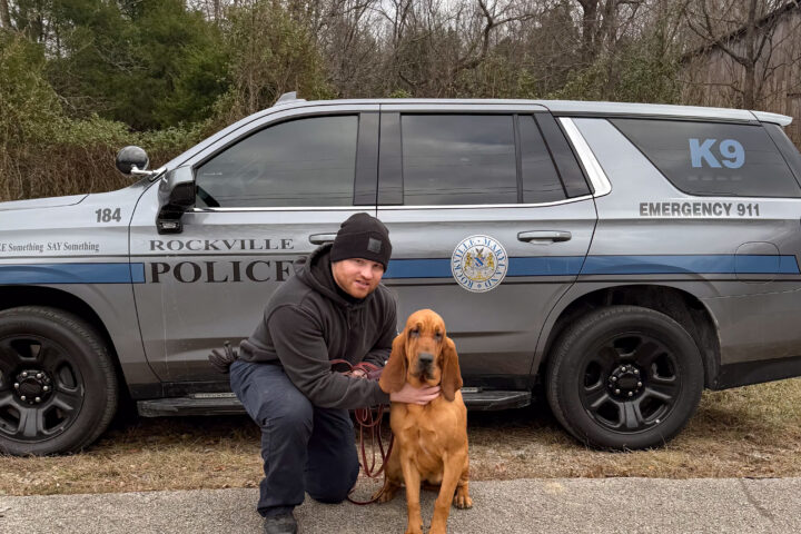 A police officer kneels next to a bloodhound in front of a Rockville City Police K9 SUV parked by a wooded area.