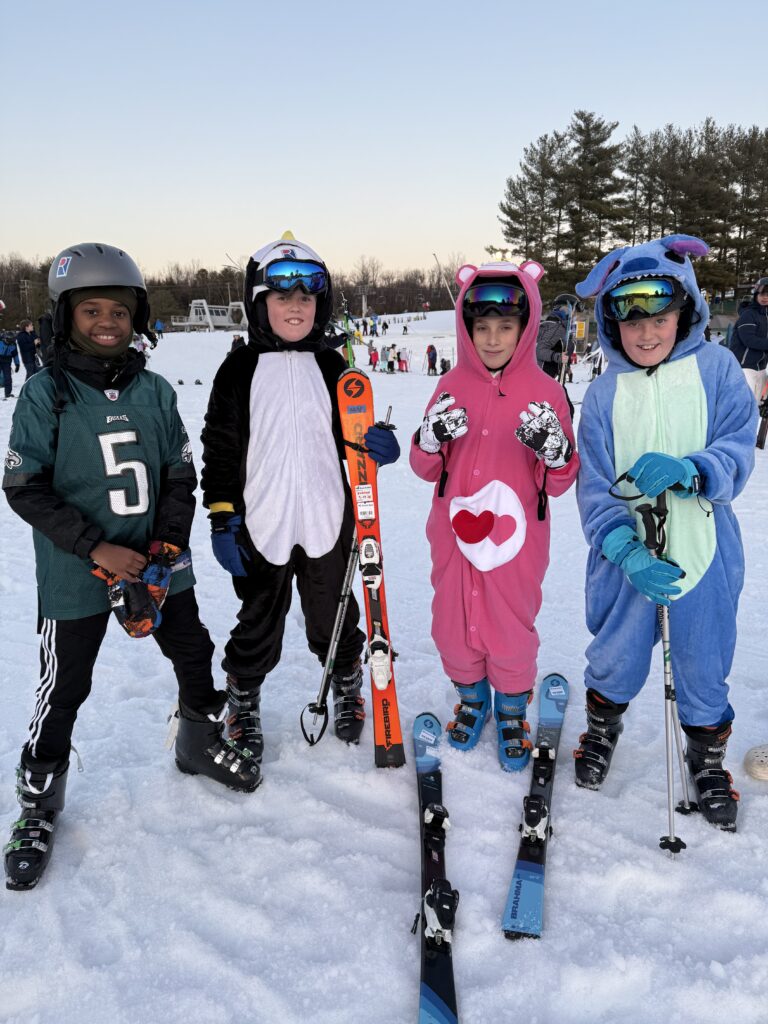 Four children stand on a snowy ski slope, three wearing animal onesies and one in a football jersey and ski helmet, with ski equipment visible around them.