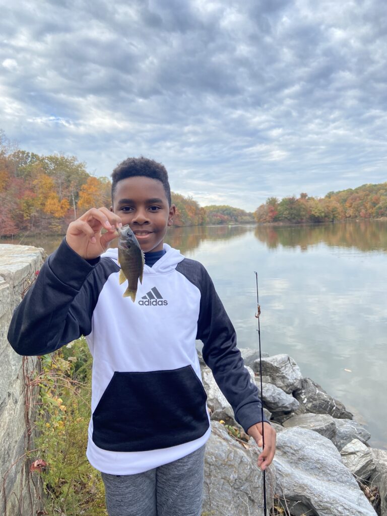 A boy in a black and white hoodie holds a small fish in one hand and a fishing rod in the other, standing by a lake with autumn trees in the background.