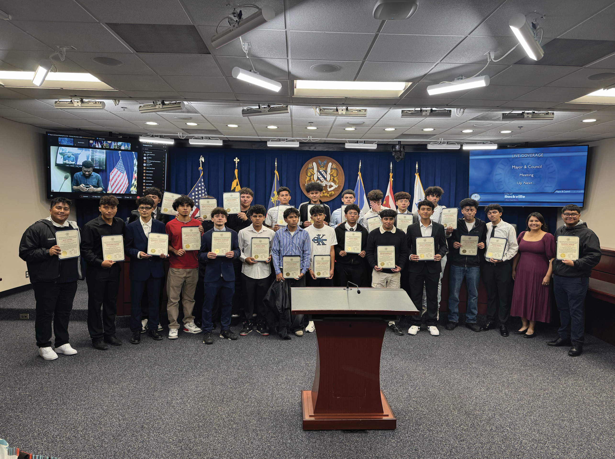 A group of young people and two adults stand in a conference room, holding certificates and posing for a photo, with flags and a podium in the background.
