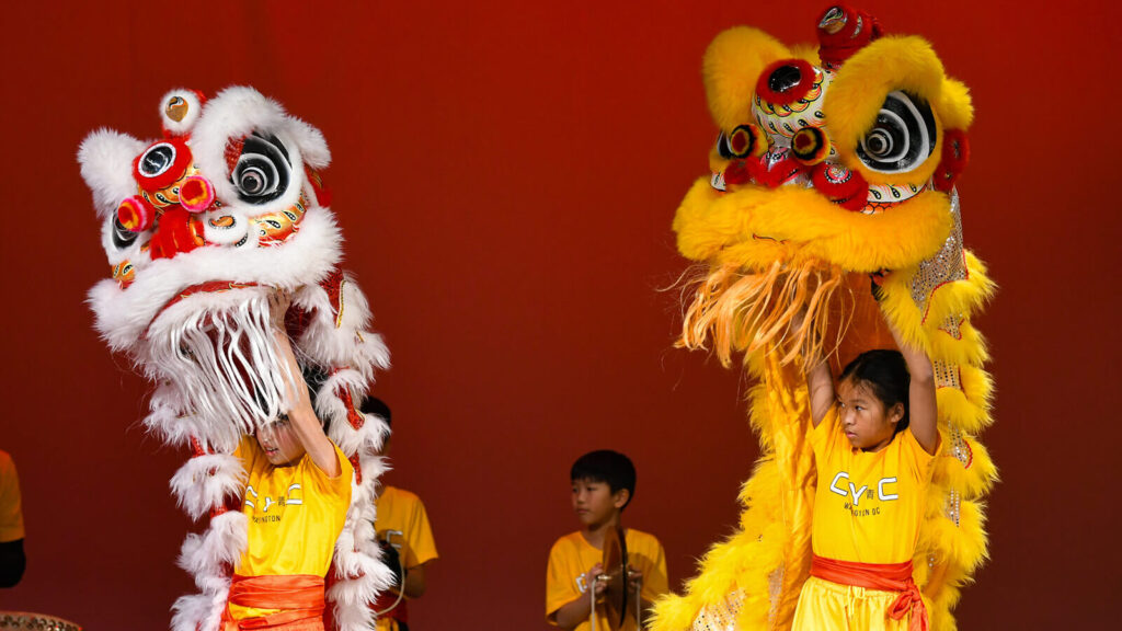 Children in yellow shirts perform a traditional lion dance on stage, holding up colorful lion costumes against a red background.
