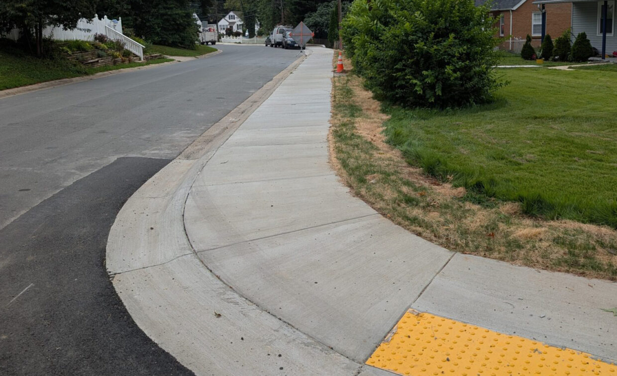 A newly paved concrete sidewalk with a yellow tactile paving section curves along a residential street with grass and houses nearby.