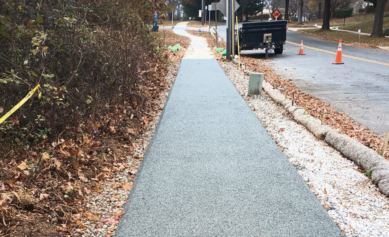 A newly constructed sidewalk runs parallel to a road with traffic cones, a trailer, and autumn leaves scattered along the edges.