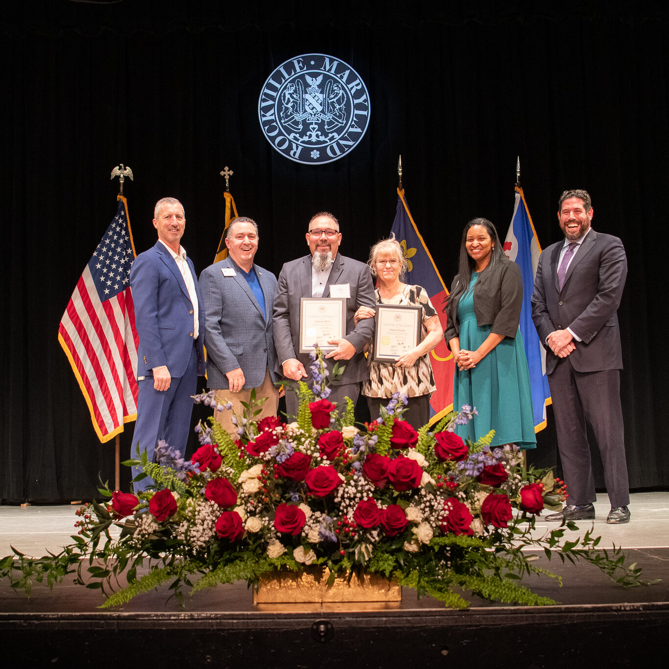 Six people stand on a stage holding framed certificates, with flowers in front and flags and a seal in the background.