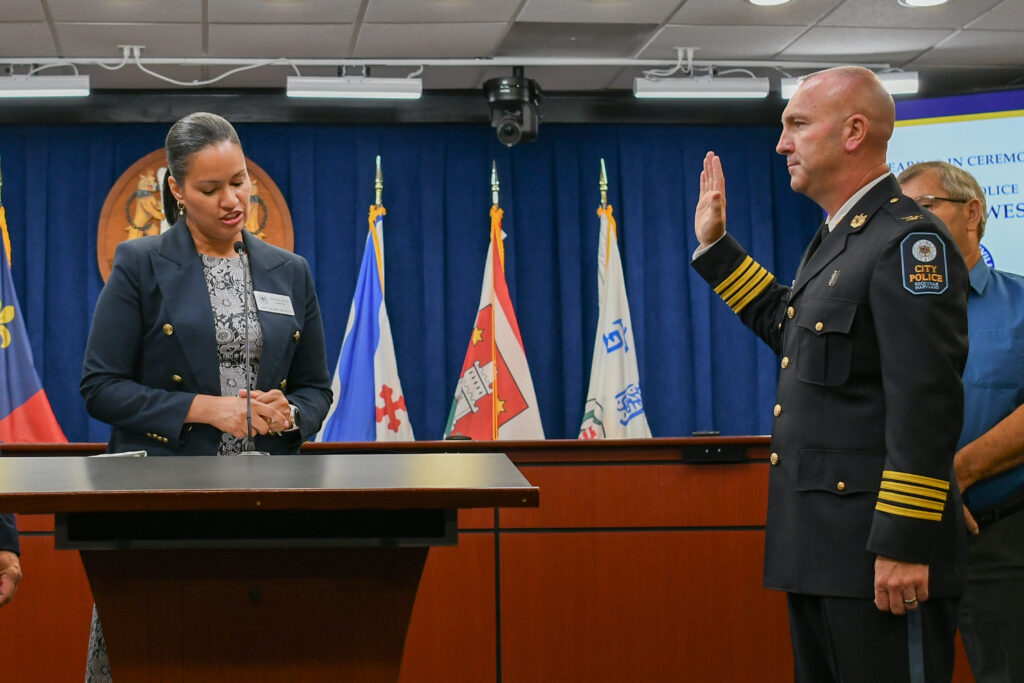 A uniformed police officer raises his right hand to be sworn in at a podium, while a woman stands next to him reading from a document. Flags are displayed in the background.
