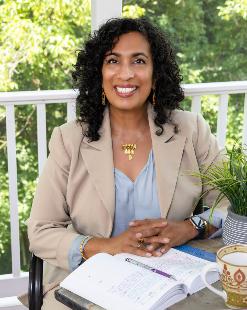 Woman with curly hair wearing a beige blazer sits at a table with an open notebook, a plant, a cup, and pen, smiling, with greenery visible in the background.