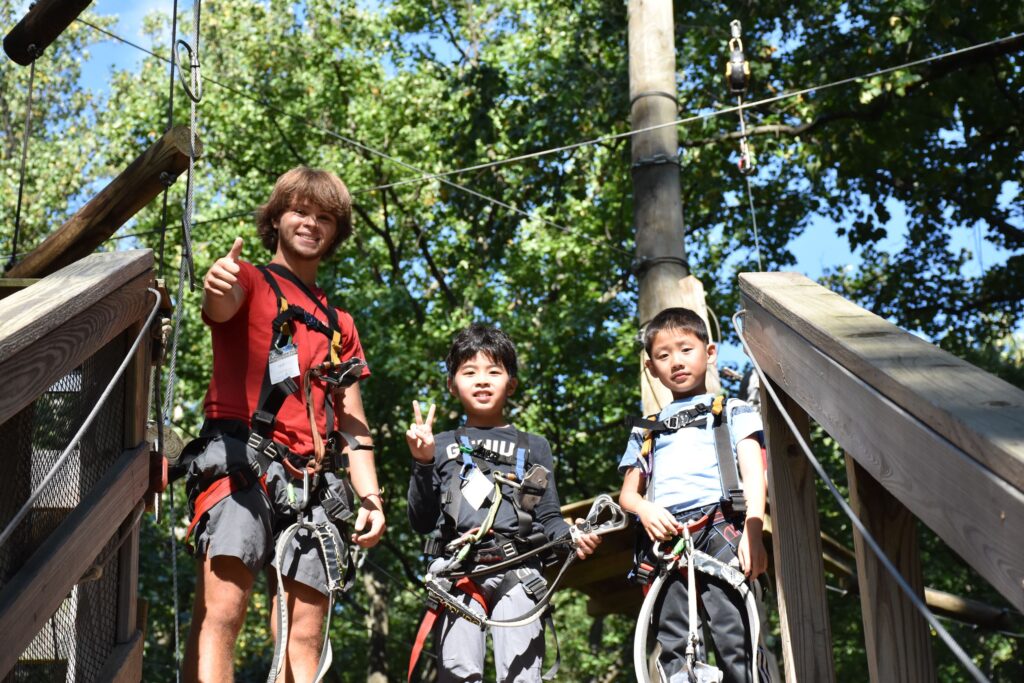 Three people wearing harnesses stand on a wooden platform outdoors, with trees and rope course equipment visible in the background.