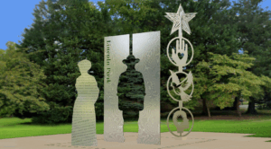 Three stainless steel sculptures stand on a paved area in a park, featuring abstract human silhouettes, engraved text, and symbolic hand and star shapes, with trees in the background.