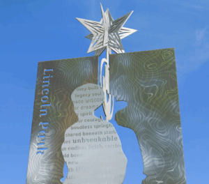 A metal sculpture in Lincoln Park features a star atop a silhouette cutout with engraved words and a textured background, set against a clear blue sky.
