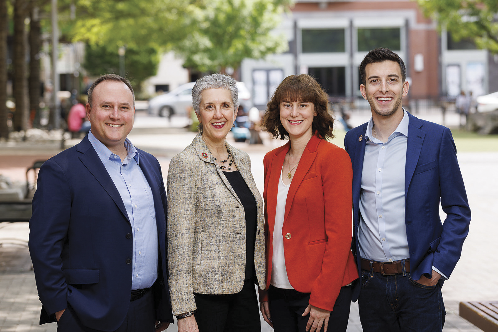 Four people in business attire stand outdoors, smiling at the camera, with trees and a building visible in the background.