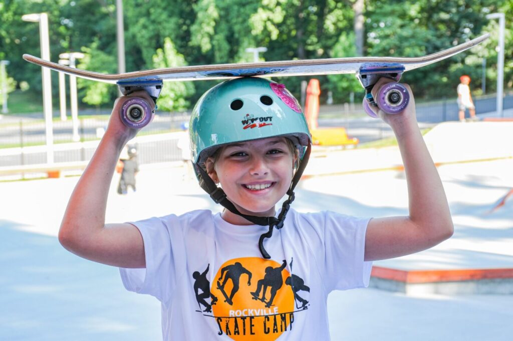 A child wearing a helmet and a "Rockville Skate Camp" T-shirt holds a skateboard overhead at an outdoor skate park.
