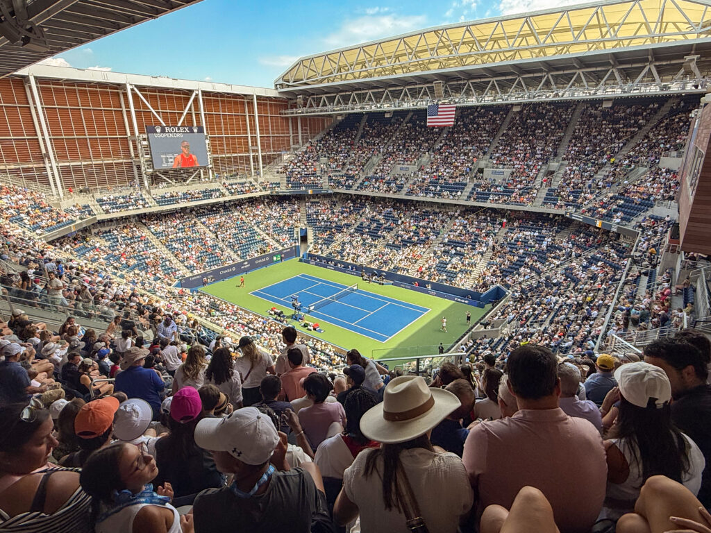 Crowd seated in a large stadium watching a tennis match on a blue court under a partially open roof during the day.