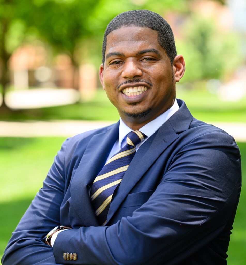A man in a navy suit and striped tie stands outdoors with arms crossed, smiling at the camera. The background is blurred greenery.