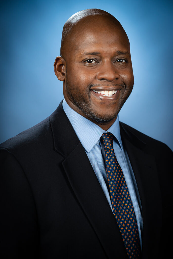 A man in a dark suit, light blue shirt, and patterned tie smiles at the camera against a blue background.