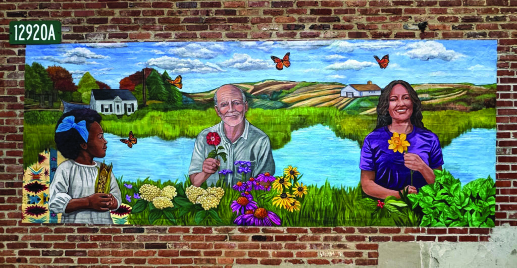 A mural on a brick wall depicts a young girl, an elderly man, and a woman holding flowers, with a scenic landscape, houses, and butterflies in the background.