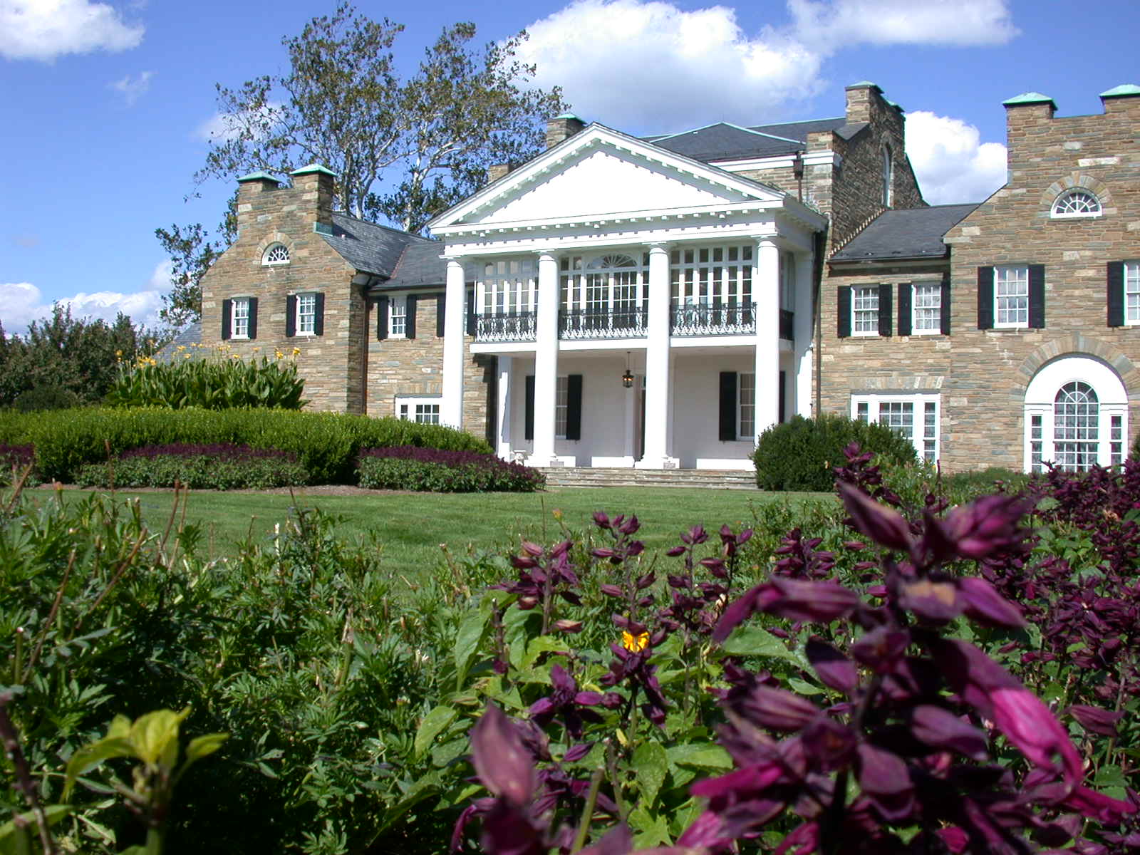 Large brick mansion with white columns and portico, surrounded by green lawns and purple flowers under a partly cloudy sky.