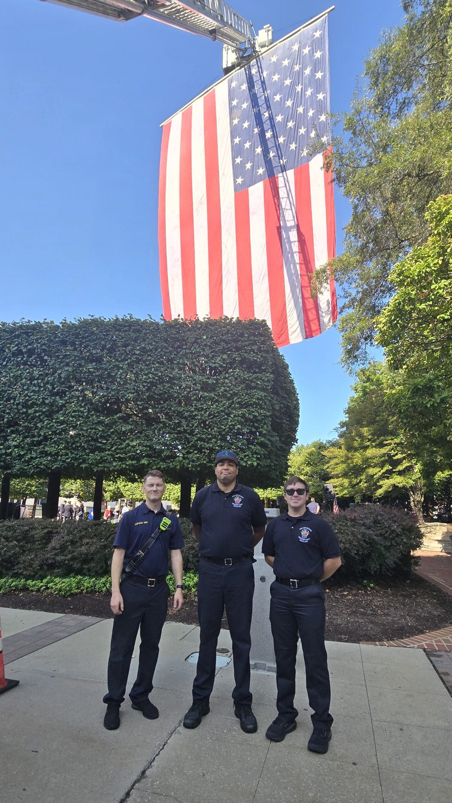 Three firefighters in uniform stand on a sidewalk in front of a large American flag hanging overhead, with trees and clear blue sky in the background.