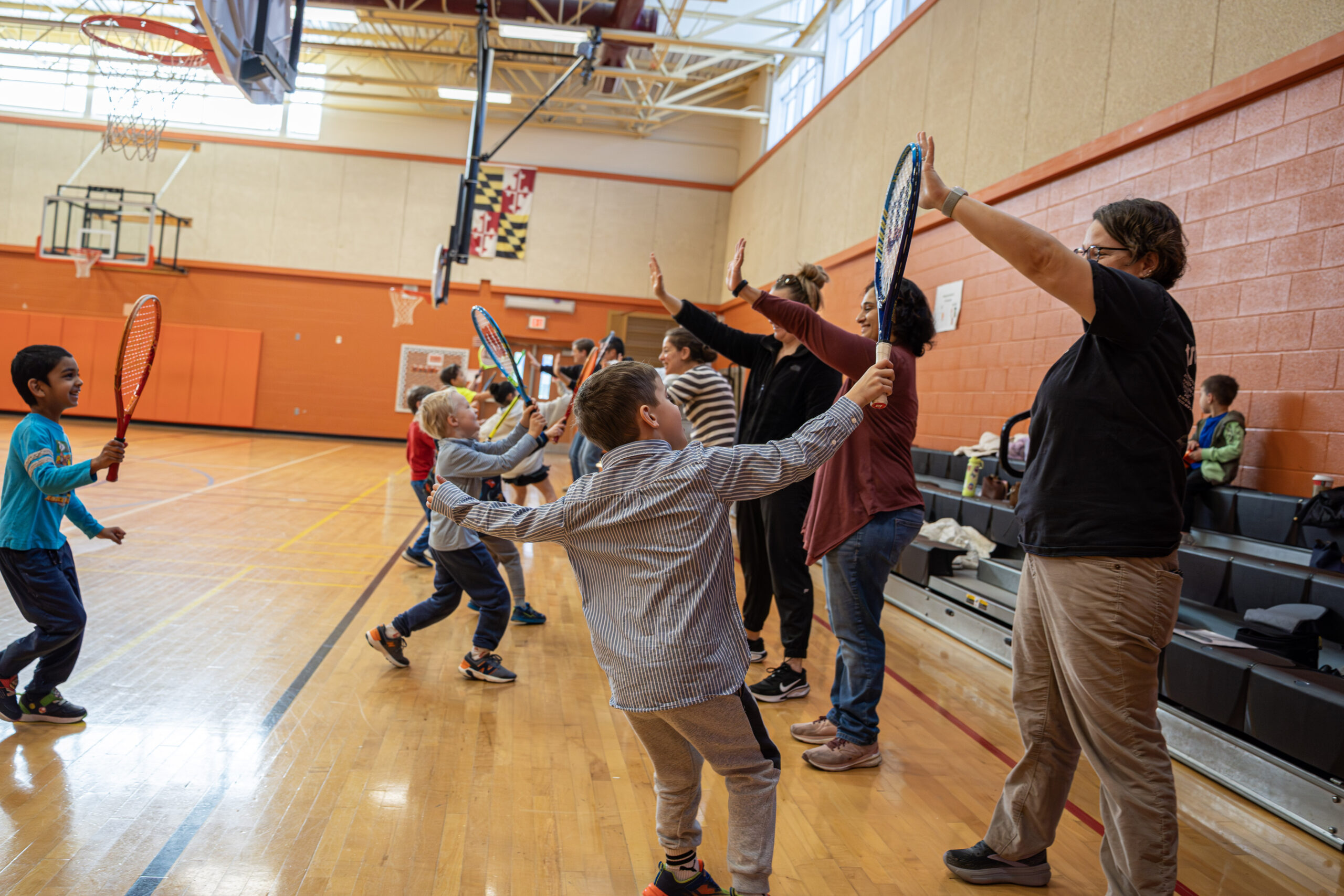 Children and adults in a gym hold tennis rackets and high-five each other while standing in two facing lines.